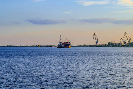 A large cargo ship departs from the Kherson port on the Dnieper river (Ukraine). Evening panorama with watercraft on the blue surface of the water and harbor cranes on the shoreの写真素材