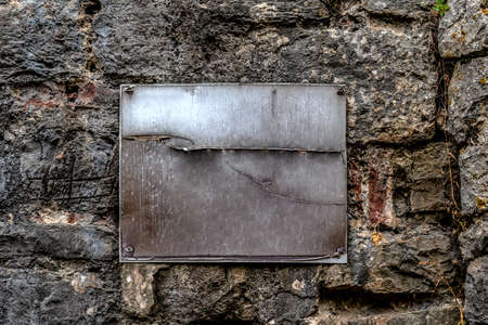 A metal rusty scratched blank plaque screwed with four screws to a stone wall. Vintage rough background with copy space. Real templateの写真素材