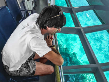 Adult young European man admiring the coral reefs of the Red Sea through the transparent glass bottom of a tourist ship. Tourist marine service in Sharm El Sheikh, Egyptの写真素材