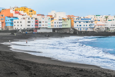 Waves wash black sand with white foam at Playa Maria Jimenez beach in Puerto de la Cruz during a storm, Spain. Several people rest in Atlantic Ocean on the backdrop of beautiful buildings in Canariesの写真素材