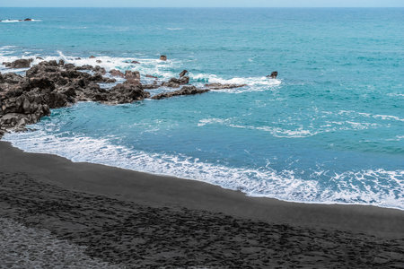 Playa Chica beach with black sand in Puerto de la Cruz in the Canary Islands, Spain. Atlantic Ocean coastline and rocks in blue raging water with white foam whipped up by wavesの写真素材