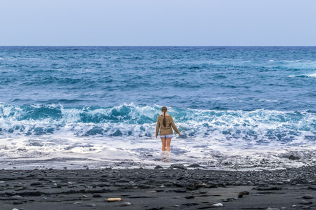 A dressed girl stands among the waves of the Atlantic Ocean on a black sand beach during a storm in the Canary Islands, Spain. Lonely young girl enjoying the sea in Tenerife in cloudy weatherの写真素材