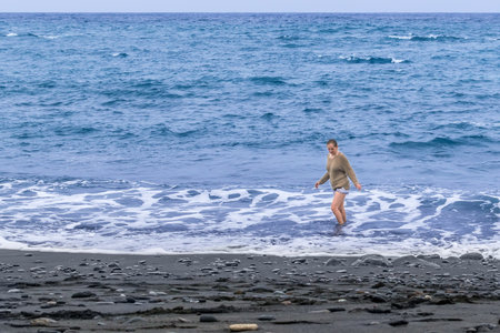 Young woman walks in the water of the Atlantic Ocean on a black sand beach in Tenerife, Spain. Lonely woman enjoying the sea on a cloudy day in the Canary Islandsの写真素材