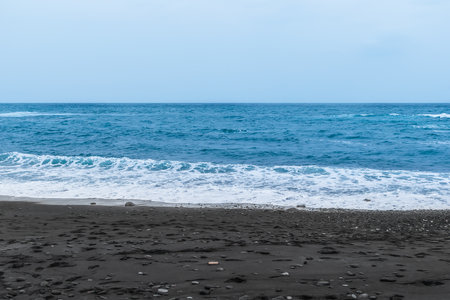 Black sand on the beach against blue turbulent water with white foam. Empty volcanic sand beach on the Atlantic Ocean in Tenerife, Canary Islands in Spainの写真素材