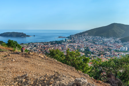 View from the top of the mountain to the city of Budva down in the valley on the Adriatic Sea, Montenegro. A young man with a backpack, squatting, looks at the seascape with Sveti Nikola Islandの写真素材