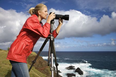 Caucasian mid-adult woman looking through camera on tripod on cliff overlooking ocean in Maui,...