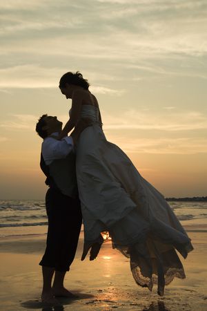 Caucasian mid-adult groom lifting up bride on beach at sunset.の写真素材