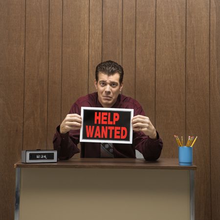 Caucasion mid-adult retro businessman with desperate expression sitting at desk holding up help wanted sign .の写真素材