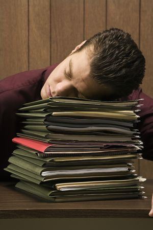 Caucasion mid-adult retro businessman sitting at desk with head down sleeping on a tall stack of...