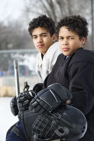 Two boys in ice hockey uniforms sitting on ice rink sidelines looking.の写真素材