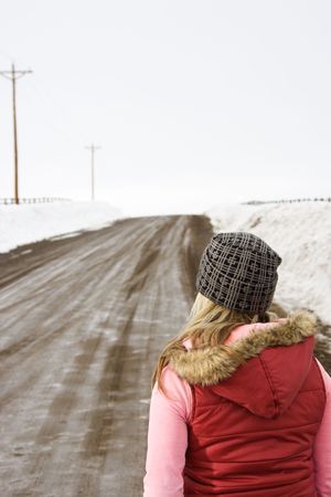 Young woman in winter clothes standing alone looking down a muddy dirt road.の写真素材