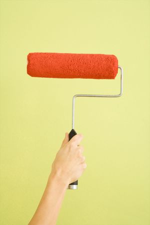 Caucasian female hand holding paint roller dipped in red against green wall.