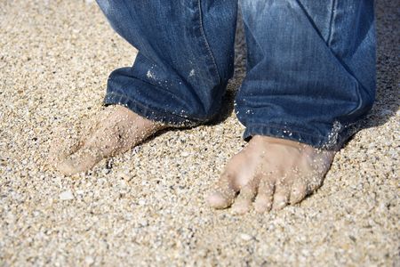 Mid-adult Caucasian male feet in jeans on sandy beach. 
