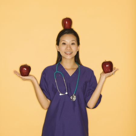 Asian Chinese mid-adult female doctor holding two red apples in hands and balancing one on head against yellow background smiling and looking at viewer.の写真素材