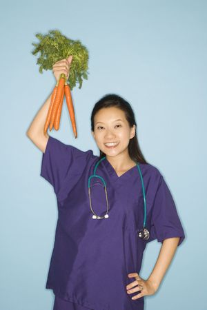 Asian Chinese mid-adult female doctor with hand on hip holding up bunch of carrots against blue background smiling and looking at viewer.の写真素材