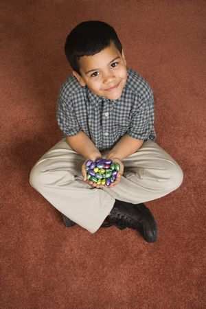 Hispanic boy sitting on floor holding chocolate candy eggs in his hands looking up at viewer smiling.の写真素材