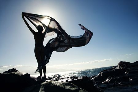 Filipino young  woman standing on rocky beach sihouetted against sky holding flowing fabric.の写真素材
