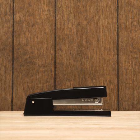 Black stapler on desk with wooden paneling.の写真素材
