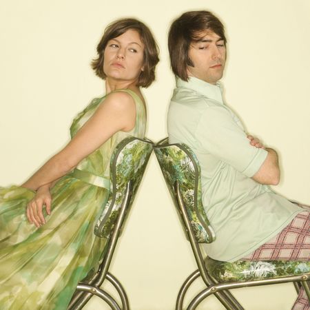 Caucasian mid-adult couple wearing vintage clothing sitting back to back in green vinyl chairs with arms crossed looking angry.の写真素材