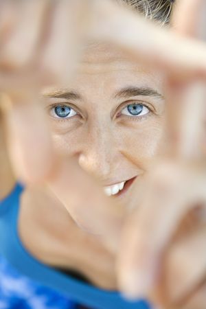 Close-up of Caucasian mid-adult woman smiling and looking at viewer through framed fingers.