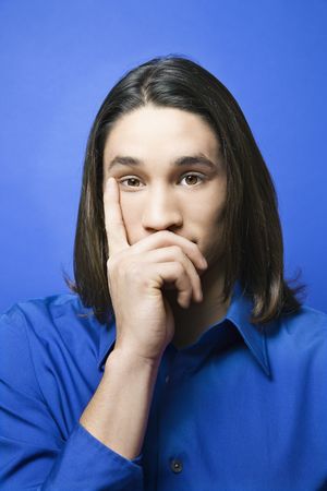 Portrait of Asian-American teen boy with hand covering chin and finger on side of face against blue...