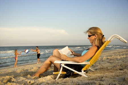 Woman at the beach reads a book while her family plays in the background by the ocean.の写真素材