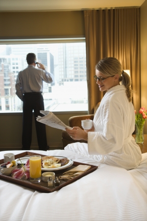 Caucasian woman in a robe sits on a hotel bed while reading the newspaper. A man stands in the background talking on his mobile phone. Vertical shot.の写真素材