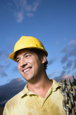 Attractive Caucasian construction worker in a yellow hardhat carries a heavy chain over his shoulder...