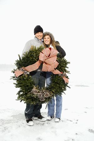 Young adult couple in the winter holding a Christmas wreath and with arms around one another. Vertical shot.の写真素材