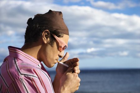 Close-up of a young man standing by the ocean and lighting a cigarette. Horizontal shot.の写真素材