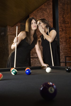 Two attractive young women at pool table as one whispers into other's ear.の写真素材