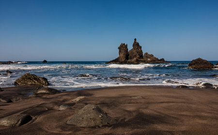 Rocky beach with black sand and ocean waves under a clear blue sky.の写真素材