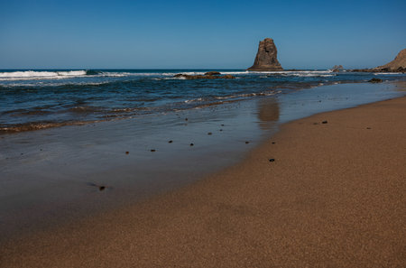 Serene beach with calm waves and a towering rock formation under a clear blue sky.の写真素材