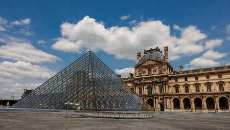 The Louvre Museum with its iconic glass pyramid under a bright blue sky with scattered clouds.の写真素材