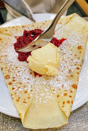 Sweet crepe with vanilla ice cream raspberry sauce and powdered sugar served on a white plate.