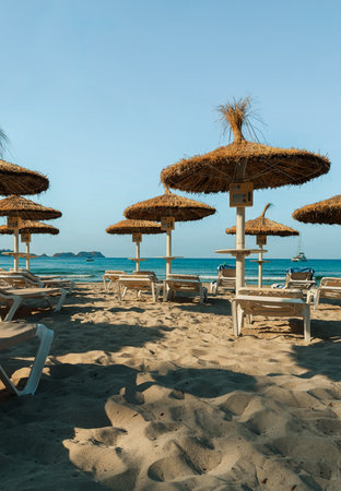 Sunny beach with straw umbrellas and empty lounge chairs facing turquoise sea.