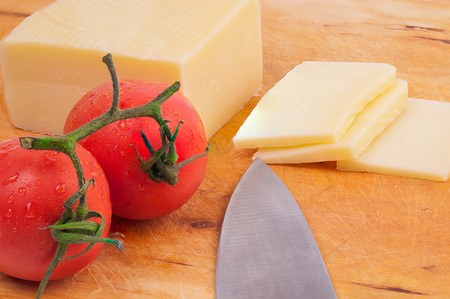 Piece of cheese, slices of cheese and a knife with  tomatoes on a used wodden boardの写真素材