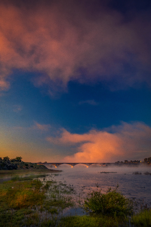 Dramatic clouds over a road bridge above a river on a summer morning with some fog above the waterの写真素材