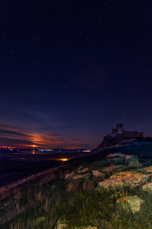 Ancient citadel found in Romania visited by tourists on top of a rocky hill during the night on a starry sky with the moon setting behind some clouds with rich rock formations in the foregroundの写真素材