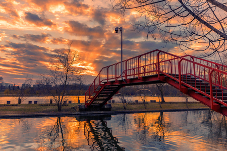 Sunset seen from a park in Bucharest with sun rays coming out from the clouds and a red metal bridge in the foreground shot at the beginning of the springの写真素材