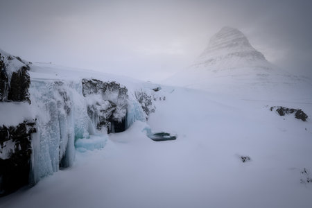 The iconic Kirkjufell mountain in Iceland, in the Snaefellsnes peninsula, famous location, winter on a very moody day with low clouds and fog, waterfall frozen with iciclesの写真素材