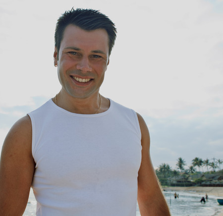 young brunette man on beach at sea smiling, gomosexual in white dress big muscles close upの写真素材