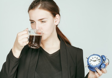 young beauty woman in business style costume waking up for work early morning on white background with clock dreanking coffeeの写真素材
