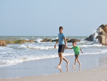 happy family on beach playing, father with son walking sea coast, rocks behind smiling enjoy summer vacationの写真素材