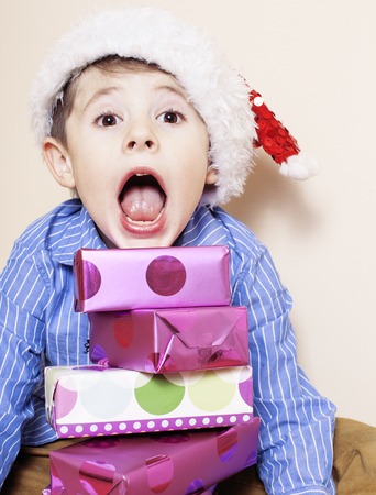 little cute boy with Christmas gifts at home. close up emotional face on boxes in santas red hat. mischief. adorableの写真素材