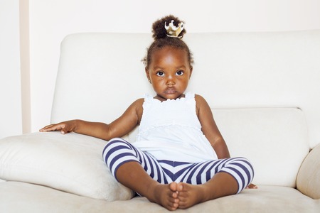 little pretty african american girl sitting in white chair wearing toy crown on head like princess or queen, lifestyle people concept close upの写真素材