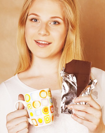 young cute blond girl eating chocolate and drinking coffee close up on warm backgroundの写真素材