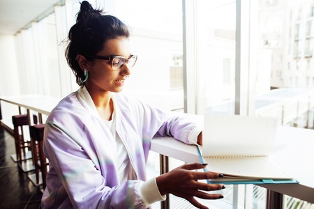 young cute hipster girl student sitting in cafe with notebook reading, wearing glasses, lifestyle happy smiling people concept close upの写真素材