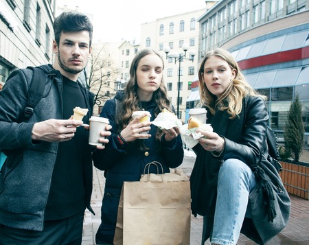lifestyle and people concept: two girls and guy eating fast food on city street together having fun,...