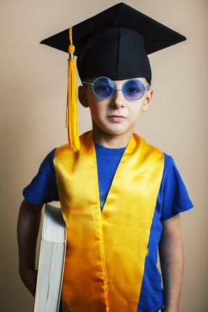 little cute preschooler boy in glasses with books at home education in graduate hat smiling posing...